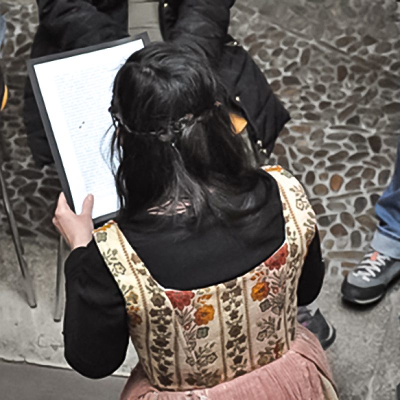 Mujer de espaldas leyendo e interpretando un texto con público al fondo